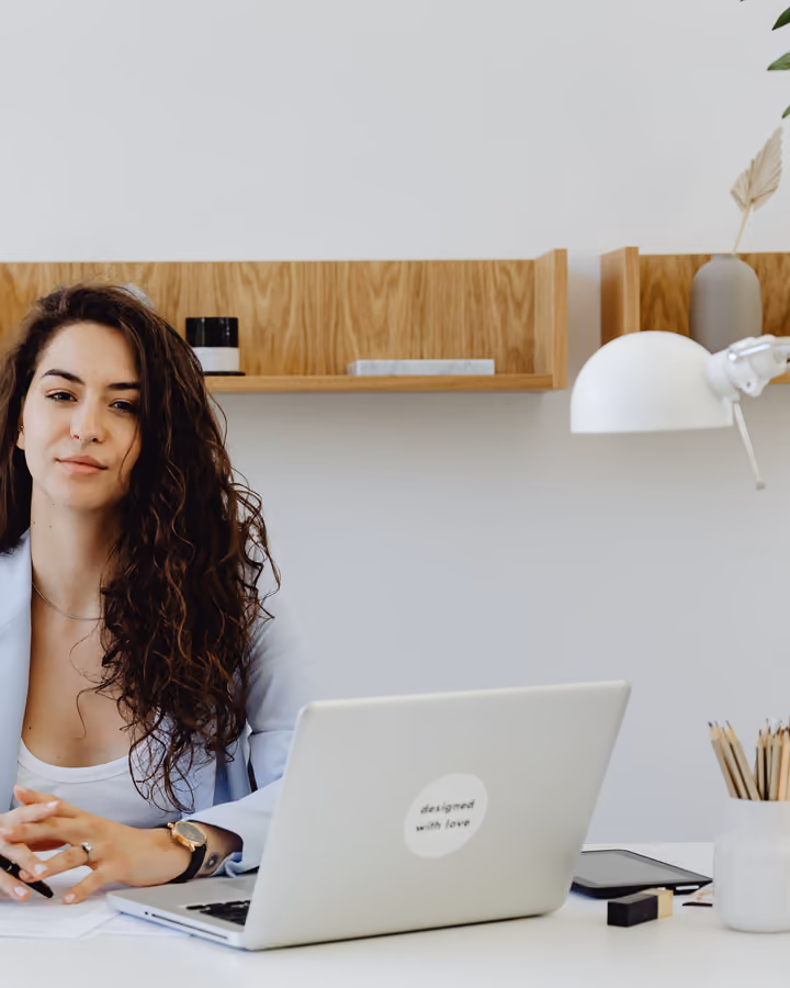 Mulher de cabelo cacheado e blazer claro sentada à mesa, com notebook, olhando para a câmera.