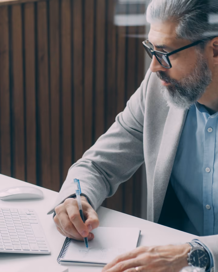 Homem de cabelo grisalho, barba e óculos escrevendo num caderno, com paletó claro e camisa azul.