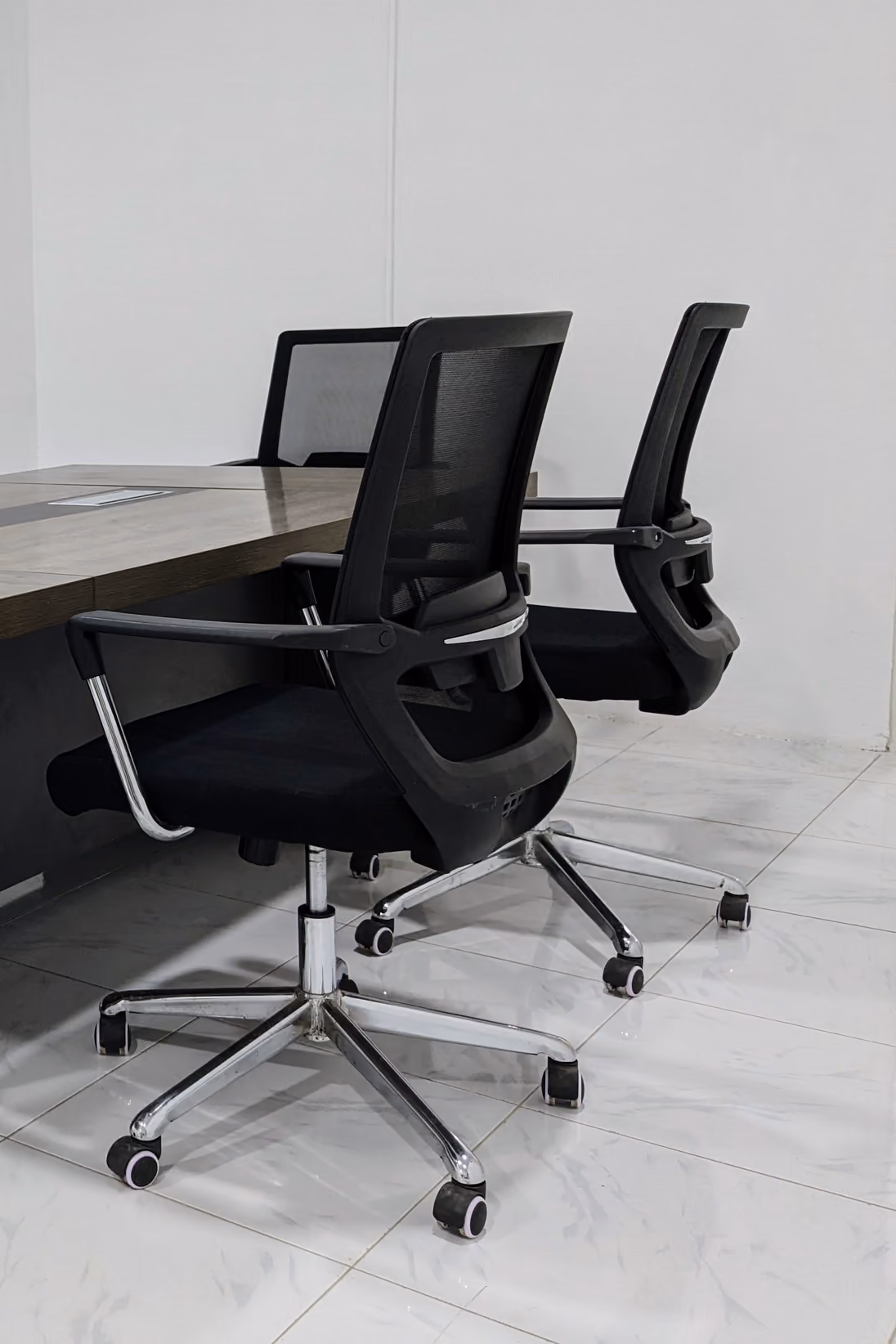 Duas cadeiras de escritório pretas, com encosto de tela, junto a uma mesa de reunião de madeira, sobre piso de porcelanato claro.